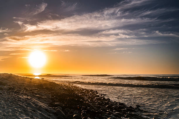 Golden sunset shines over a beach with ocean waves gently rolling onto a rocky shoreline under a sky with wispy clouds