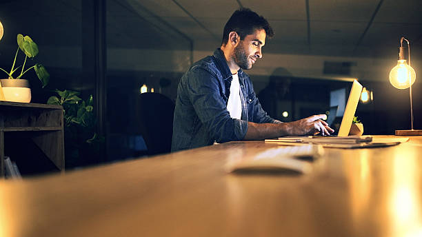 istock Laptop, night and typing with business man in office for deadline or feedback on creative report. Computer, desk and research with design employee in dark workplace for productivity at zero hour 2224204921