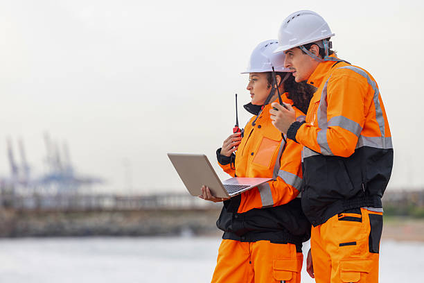 istock Dock Worker in Safety Uniforms Using Technology for Port Operations, Maritime Safety Engineers Working at Harbor Site, Industrial Engineers Collaborating at Port Terminal with Laptop and Walkie-Talkie 2224221028