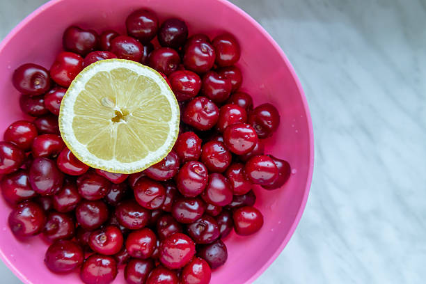 Freshly washed red cherries in a pink plastic bowl with a slice of lemon placed on top. The bright colors and clean fruit suggest freshness, healthy eating, and summer flavors.