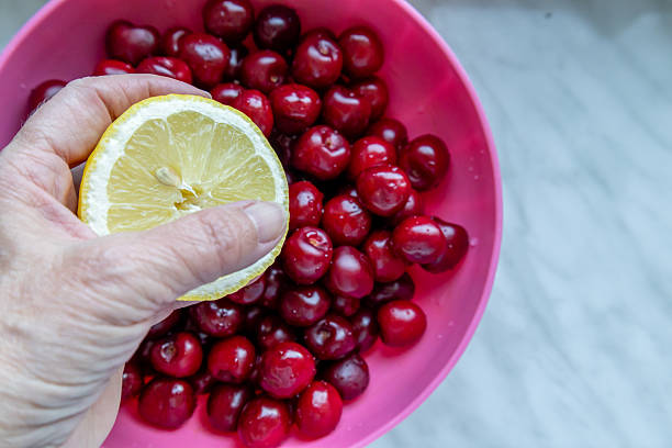 A hand holding a slice of lemon above freshly washed red cherries in a pink plastic bowl. The composition suggests fruit preparation, healthy eating, and the refreshing flavors of summer.