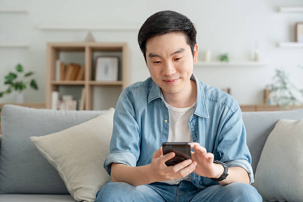 istock Smiling handsome young Asian man sitting on cozy couch, using funny mobile apps in living room. man at home, doing online shopping, messaging friends, posting on social media 2224222880
