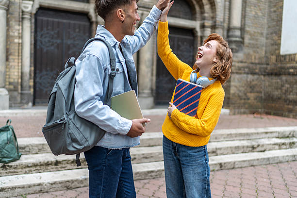 Two cheerful young adult university students are giving high five to each other while celebrating success in front of their university building