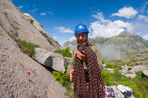 Bearded man in blue helmet coiling climbing rope on rocky mountain terrain. Outdoor adventure enthusiast readying gear for rock climbing. Mountain sports and safety preparation.