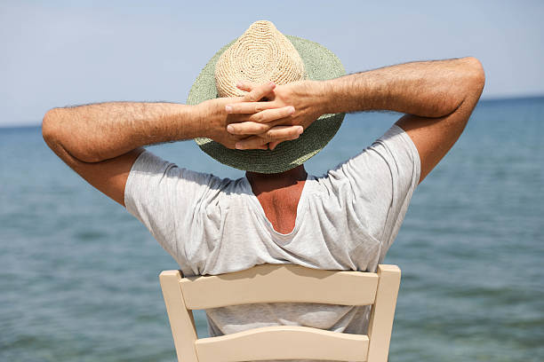 Rear View Of A Senior Man Sitting In A Chair On The Beach, Wearing A Straw Hat And Relaxing With Hands Behind Head While Looking At The Sea. Tranquil Summer Vacation Concept, Outdoor Leisure By The Ocean.