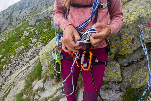 Woman wearing harness and climbing shoes adjusting ropes on rocky mountain face. Outdoor adventure with safety equipment for rock climbing. Mountain climbing and hiking activity in nature.
