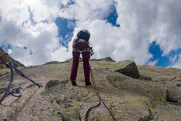 Climber wearing harness and backpack scaling a rocky cliff with safety ropes attached. Outdoor adventure sport on rugged mountain terrain. Person engaged in challenging rock climbing activity.
