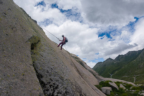 Climber wearing helmet and harness abseils down a large rock face on a mountain. Outdoor adventure sport in nature with mountainous landscape and cloudy sky background. Extreme sports activity.