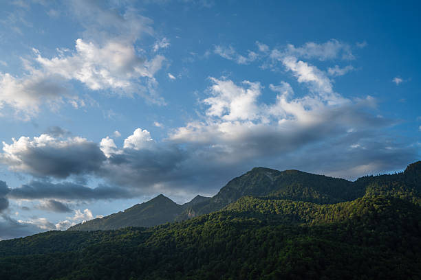 A breathtaking view of a mountain range under a clear blue sky, adorned with fluffy clouds and lush green forests. Capturing nature's tranquility and grandeur, it's a perfect depiction of scenic landscape.
