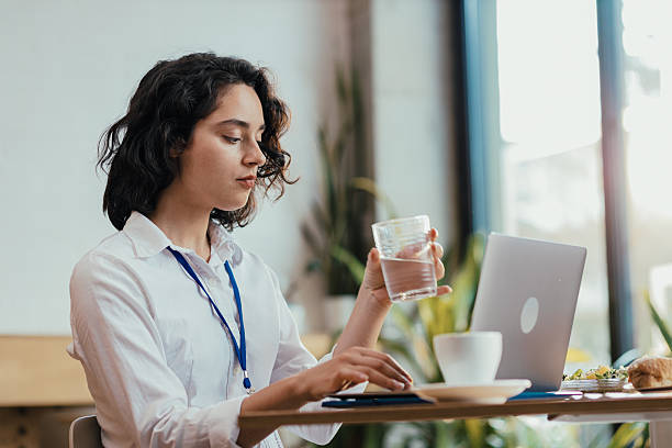 istock Young woman drinking water while typing on laptop during lunch break in bright office cafeteria 2224251801