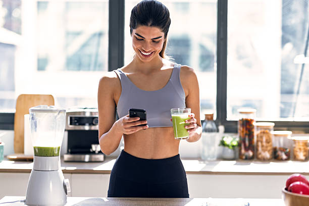 istock Sporty young woman drinking handmade a vegetable green smoothie while using smartphone in the kitchen at home. 2224254093
