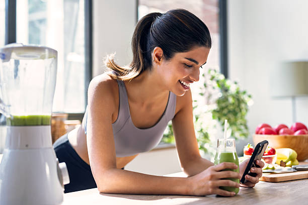 istock Sporty young woman drinking handmade a vegetable green smoothie while using smartphone in the kitchen at home. 2224254121