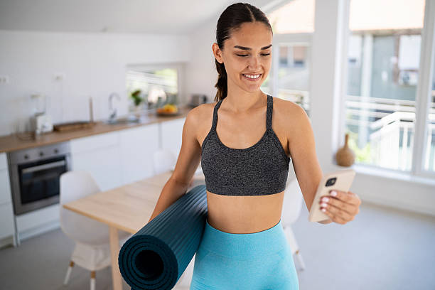 istock Young woman at home, dressed in activewear, holding a rolled-up exercise mat in one arm while looking at her smartphone. She’s either preparing for or finishing a workout session, possibly following an online fitness class or checking a workout app. 2224254145