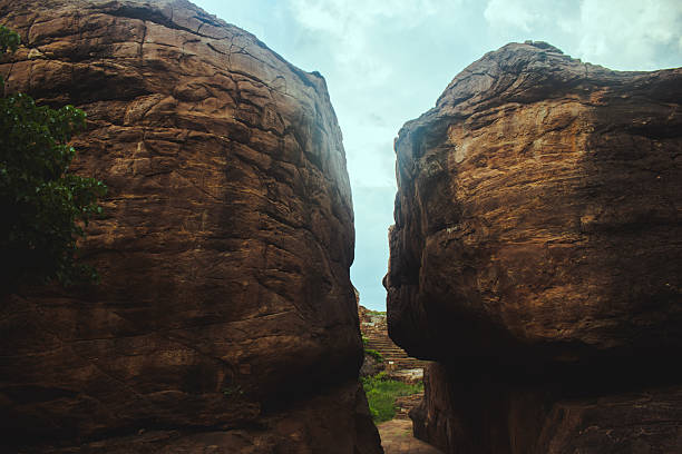 Towering reddish-brown cliffs split by a narrow crevice under a dramatic cloudy sky at Badami, karnataka, India.