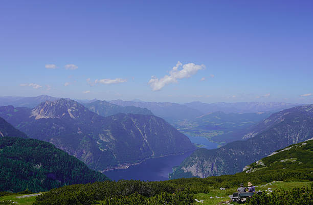 A vast, high-altitude panoramic view captured from the summit of Saukarfunktel (likely one of the 'four peaks' above Zauchensee) in the Radstädter Tauern region of Austria. The foreground features the gentle, rolling green and brown slopes of the mountain ridge, with a small access road and possibly a lift station in the middle ground, indicating high-alpine infrastructure. The background is a breathtaking display of numerous receding mountain ranges, creating layers of deep blue and gray-green peaks fading into the distance. Patches of snow are visible on the far horizon, contrasting with the soft, cloudy sky. This image captures the immense scale and layered beauty of the Austrian Alps.