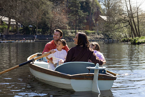 A wide shot of a cheerful family of four enjoying a relaxing rowing boat ride on a sunny day at a scenic lakeside location. The father rows the boat with a smile while the son sits on his lap, laughing. The mother and daughter face them, sharing a joyful moment. This image captures the essence of quality family time, outdoor adventure, and bonding.Videos are available similar to this scenario.