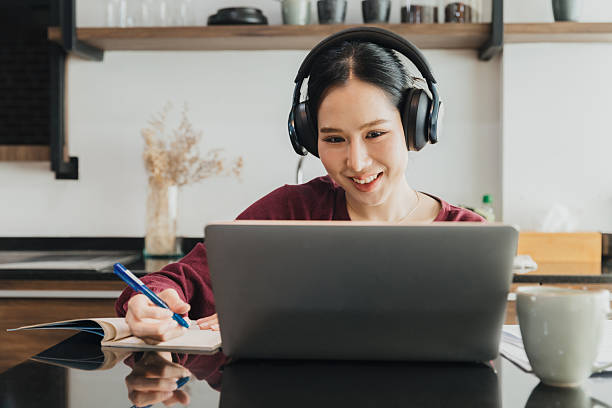istock Woman studying at home using a laptop while wearing headphones and taking notes in a cozy kitchen setting 2224272478