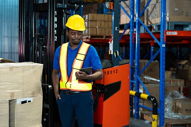 African male adult warehouse employee checking data on tablet standing next to pallet stack wearing safety uniform during stock monitoring inside organized logistics storage area