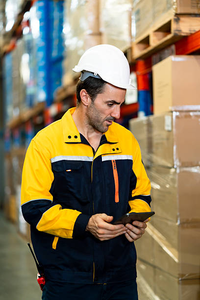 istock White adult male warehouse worker smiling while checking tablet and walking along packed boxes and shelf racks inside organized logistics facility wearing hard hat, inside logistics warehouse. 2224284381