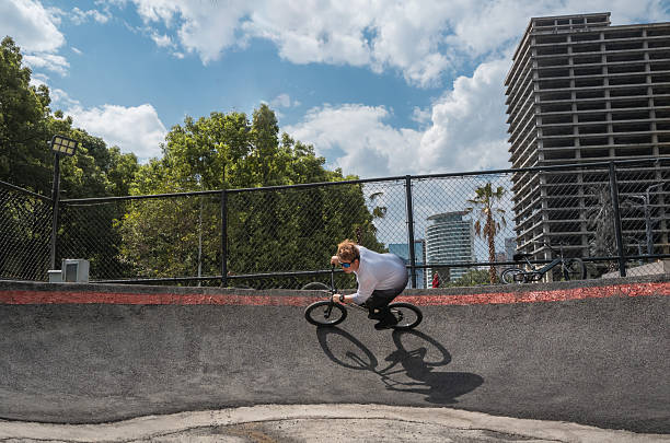 Action-packed moment capturing a BMX rider executing an impressive jump over a chain-link fence, set against a backdrop of lush greenery and partly cloudy skies. The dynamic scene conveys excitement and skill, suitable for sports, extreme sports, or action-themed content.