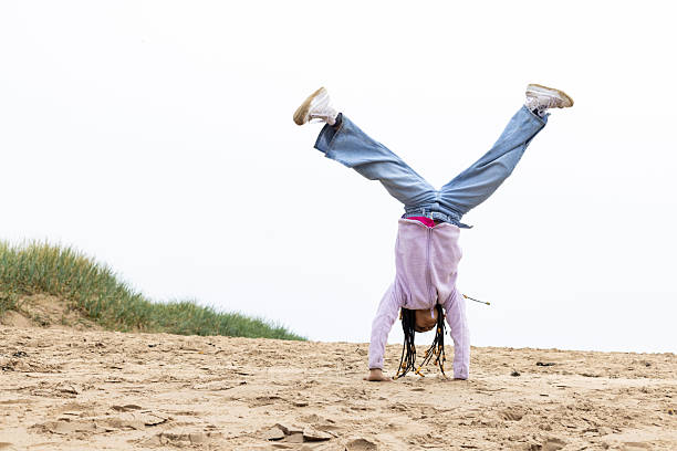 A medium wide-angle view of a young girl practising her gymnastic skills as she plays on the beach with her family in Alnmouth on the coast of Northumberland. She is doing a cartwheel on the hard sand along the beach and showing her family her flexibility.