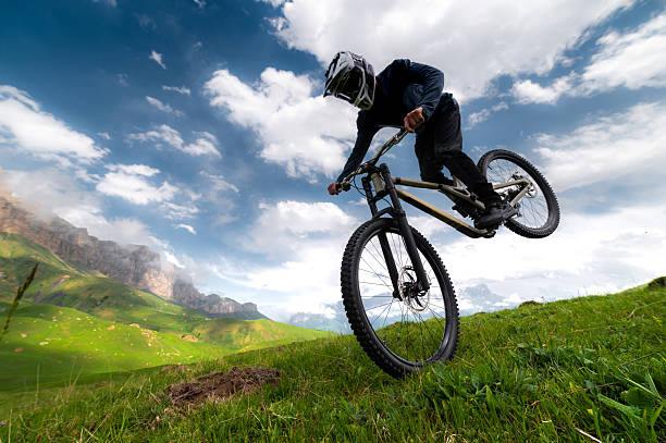 A mountain biker takes off with his rear wheel leaning on his front wheel, performing a manuel or wheelie trick, against a mountain landscape in the fields in summer.