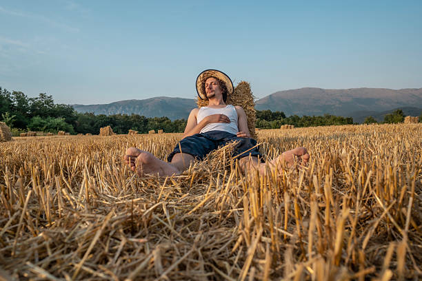 A man enjoying a serene moment seated in a harvested field during a warm summer day, with hay bales, a straw hat, and mountains in the background.