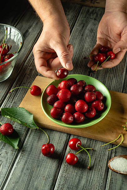 istock Hands delicately select ripe cherries from a green bowl filled with fruit in a cozy kitchen. The setting features wooden surfaces and scattered fruit, evoking a homemade feel 2224313717