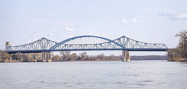 A scenic view of a steel arch bridge spanning a calm river under a clear sky, with bare trees lining the riverbanks.