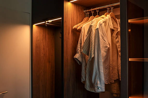 istock Modern wooden wardrobe with LED lighting and several light shirts on hangers. Minimalist interior design with open closet and contemporary door 2224384293