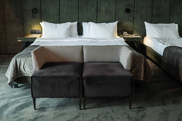 Modern hotel room interior with two beds, white pillows, gray blankets and small black sofa at foot of bed. Cozy minimalist design with soft lighting and green headboard.