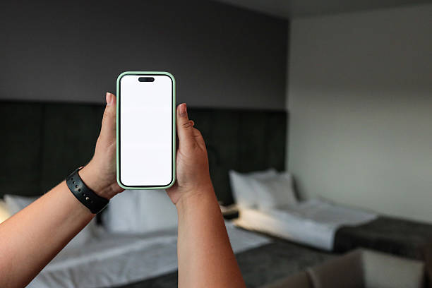 istock Person holding smartphone with blank white screen in modern hotel room. Mobile device with empty display for mockup or app presentation. Cozy interior with beds in background 2224385076