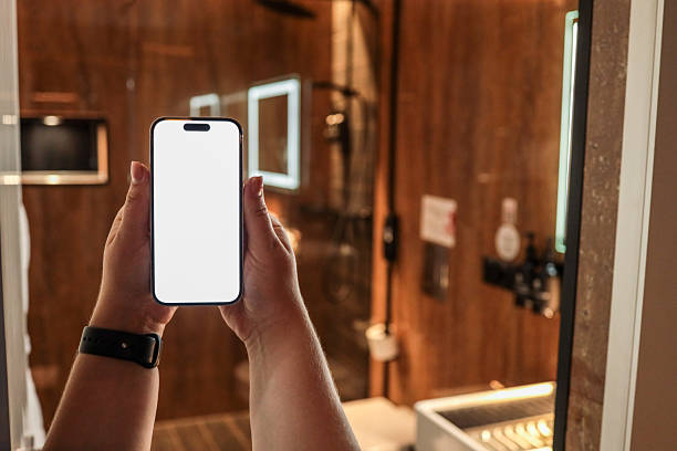 istock Person holding smartphone with blank white screen in modern hotel bathroom. Empty display for mockup, app design or advertising. Stylish interior with sink and shower in background 2224385079