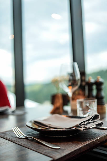 istock Close-up of elegant restaurant table setting with plate, napkin, fork, and glass in front of large window with blurred mountain view. Cozy dining interior with natural light 2224385101