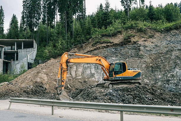 istock Excavator working at construction site near concrete building structure. Heavy machinery digging on rocky slope by the road with forest trees in background 2224385981