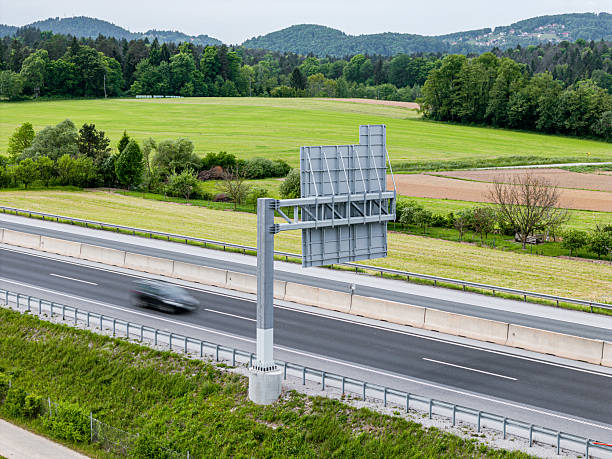 A highway scene featuring a large blank billboard beside a multi-lane road. The landscape includes green fields and trees in the background, with a blurred car passing by, indicating motion.