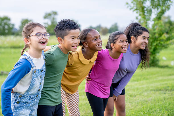 A small group of multi-ethnic school aged children huddle together outside as they pose for a portrait.  They are each dressed casually as they laugh and smile in the warm summer air.