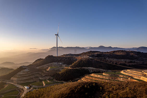 The image captures several wind turbines rising from layered terraced fields and surrounding forest on a mountain top during dusk. The setting sun casts a soft, golden glow across the landscape, highlighting the curves of the terraces and the turning blades of the turbines. The scene blends natural beauty with clean energy technology, creating a peaceful and inspiring atmosphere. This photo is suitable for categories such as renewable energy, sustainable development, rural landscapes, environmental protection, and clean technology.