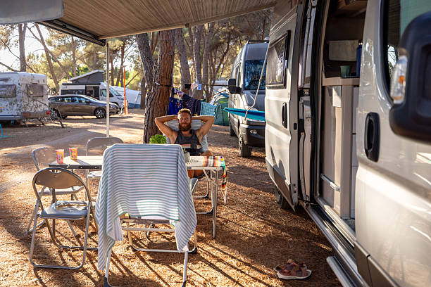 Bearded man sitting relaxed under camper awning at forest campsite. Casual outdoor leisure with camping gear and drying clothes nearby. Camping lifestyle and nature retreat.