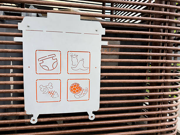 Stylized waste bin sign with minimalist icons illustrating prohibited trash types. Modern metal plate design with orange line art symbols mounted on a metal slat surface in an urban setting