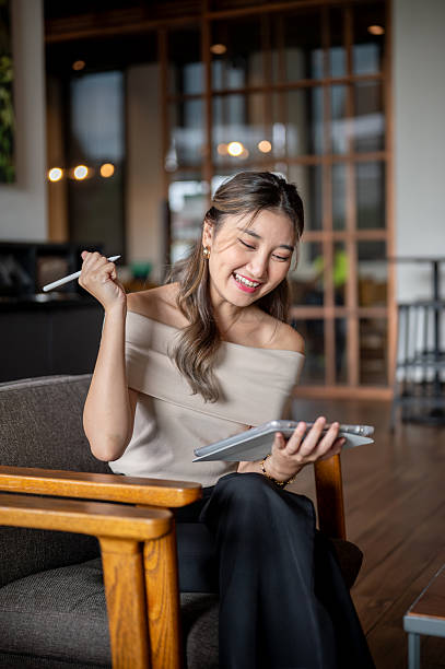 Asian woman smiling broadly while sitting and holding tablet on wooden chair in cafe or coffee shop.