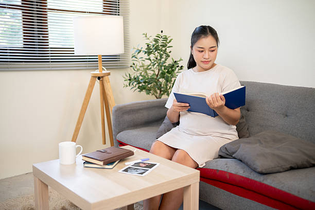istock Pregnant asian woman reading a book for her baby's brain development while sitting on sofa in house. 2224435821