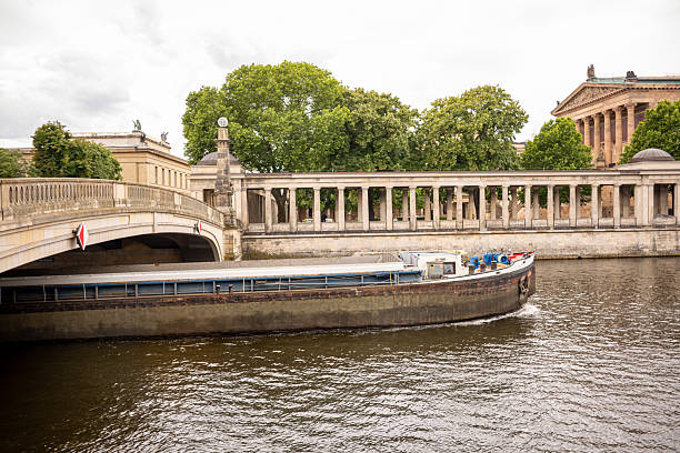 An old cargo barge navigates a calm river beside a stone bridge and colonnade. Classical architecture and water blend into a timeless scene.