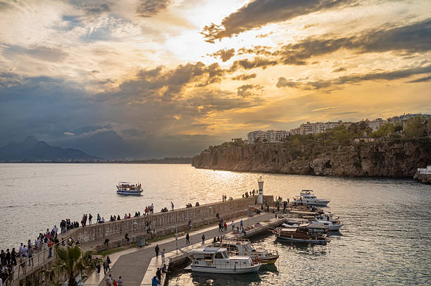 People sitting and walking on the harbour wall at dusk in Antalya, Antalya Province, Turkey (Turkiye), as a pleasure boat returns and the sky turns orange during sunset with the Taurus Mountains (Toros Mountains) visible in the distance. The calm and peaceful scene captures the beauty of Antalya's old city, known as Kaleici, which is surrounded by stunning landscapes and rich history dating back to ancient times. The old city boasts an impressive mix of Roman, Byzantine, and Ottoman architecture, making it a popular destination for tourists looking to explore Turkey's vibrant culture and heritage