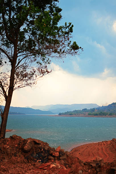 A vivid turquoise reservoir stretches between rust-colored clay banks beneath a pale, cloud-flecked sky. Branches and foliage in the foreground frame the scene, while hazy mountain ridges in the distance. Ideal for themes of landscape, travel, nature, water conservation, and dramatic outdoor vistas.