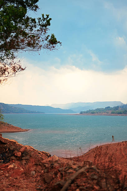 A vivid turquoise reservoir stretches between rust-colored clay banks beneath a pale, cloud-flecked sky. Branches and foliage in the foreground frame the scene, while hazy mountain ridges in the distance. Ideal for themes of landscape, travel, nature, water conservation, and dramatic outdoor vistas.