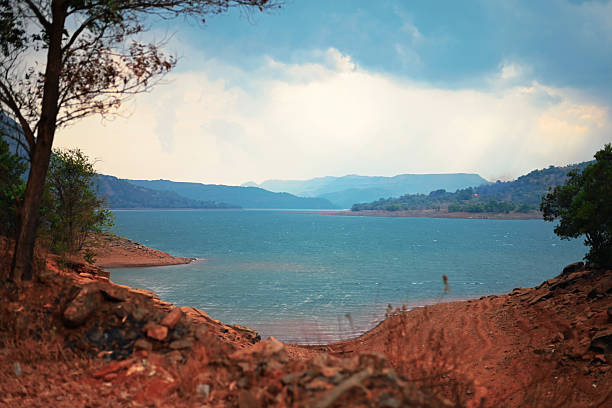 A vivid turquoise reservoir stretches between rust-colored clay banks beneath a pale, cloud-flecked sky. Branches and foliage in the foreground frame the scene, while hazy mountain ridges in the distance. Ideal for themes of landscape, travel, nature, water conservation, and dramatic outdoor vistas.