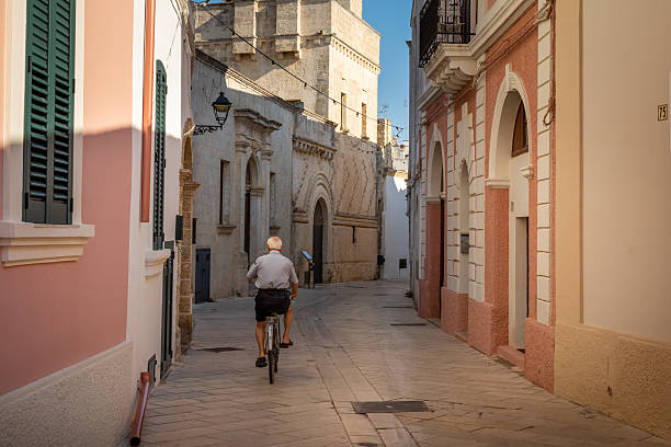 An elderly man with white hair rides a bicycle past buildings and through the old streets of Presicce, Lecce Province, Apulia, Italy.