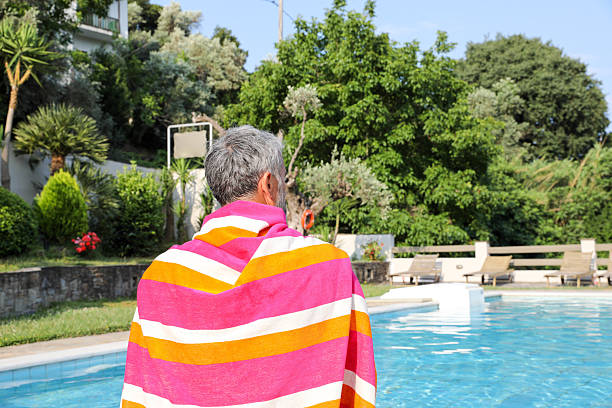 Back View Of A Senior Man With Gray Hair Sitting By A Pool, Wrapped In A Bright Striped Towel. The Scene Captures A Relaxing Moment In The Sun After Swimming, With Greenery And Lounge Chairs In The Background. Concept Of Rest, Wellness, And Healthy Aging.