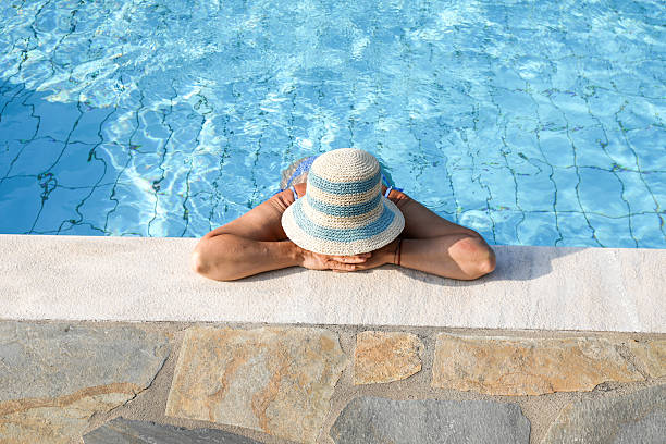 Mature Woman In Swimwear Leaning On The Edge Of A Pool With Her Arms Crossed And A Straw Hat Covering Her Head. Peaceful Summer Moment Of Relaxation And Privacy On Vacation.